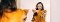 Frau in orangefarbenem Pullover mit schwarzen und weißen Streifen an den Ärmeln zeigt lächelnd mit ausgestrecktem Arm auf ihr Spiegelbild