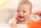 Lachendes Baby mit Essensresten im Gesicht vor einem orangefarbenen Löffel und einer orangefarbenen Schüssel