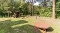Spielplatz mit Wippe, Kletterturm mit Rutsche und rundem Spielgerät auf Federn in einem bewaldeten Park