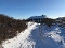 Schneebedeckter Weg zwischen niedrigen Büschen führt zu einem langgestreckten Haus unter klarem Himmel