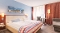 Hotelzimmer mit Doppelbett, zwei Nachttischen mit Lampen, einem kleinen Tisch mit Stuhl und einem Sessel vor einem Fenster mit Vorhängen