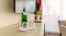 Grüner Glasflasche mit Etikett und zwei umgedrehten Gläsern auf einem hellen Tisch vor einem Fernseher und einem Fenster mit weißen Gardinen und rotem Vorhang.