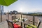 Terrasse mit Tisch, vier Stühlen mit Kissen und zwei Tassen unter grünem Sonnenschirm mit Blick auf gepflasterten Platz und Häuser im Hintergrund