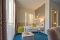 Einzelzimmer mit Bett, grünem Kissen und Decke, blauem Teppich, Nachttisch mit Lampe und kleinem Tisch mit Wasserflasche und Glas