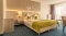 Hotelzimmer mit Doppelbett, zwei grünen Kissen und grünem Bettüberwurf, zwei Nachttischen mit Lampen, Fenster mit Vorhängen und Stuhl