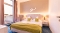 Hotelzimmer mit Doppelbett, zwei grünen Kissen, grüner Decke, zwei Nachttischen mit Lampen und Balkon mit Blick auf Dächer