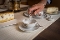Hand mit Zange hält einen Zuckerwürfel über eine weiße Tasse mit floralem Muster auf einer Untertasse auf einem gedeckten Tisch mit Kuchen im Hintergrund