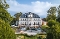 Großes historisches Herrenhaus mit Mansarddach, umgeben von Gartenanlagen und Bäumen an einem sonnigen Tag