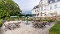 Außenansicht eines historischen Gebäudes mit Terrasse, auf der mehrere Tische, Stühle und Sonnenschirme stehen, umgeben von Bäumen und Hecken