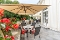 Terrasse mit Tischen, Stühlen und großen Sonnenschirmen vor einem weißen Gebäude, umgeben von grünen Bäumen und roten Blumen.