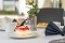Tisch auf Terrasse mit weißer Tischdecke, Teller mit Stück Erdbeerkuchen, gefalteter schwarzer Stoffserviette und unscharfem Hintergrund mit weiteren Tischen und Stühlen
