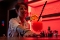 Cocktailglas mit rotem Getränk, Orangenscheibe und schwarzem Strohhalm auf Bar, unscharfe Frau im Hintergrund