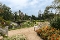 Blick auf einen Kurpark mit Kieswegen, weißen Bänken, Blumenbeeten und einem kleinen Pavillon im Hintergrund