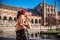 Frau mit schwarzem, besticktem Kleid und roter Blume im Haar vor einem historischen Gebäude mit Wasserbecken in Andalusien