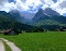 Grüne Wiese mit mehreren Holzhütten im Vordergrund und bewaldeten Bergen unter wolkigem Himmel im Hintergrund