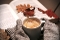 Zwei Hände in grauen Strickhandschuhen halten eine weiße Tasse mit schaumigem Kaffee vor einem aufgeschlagenen Buch mit Herbstblättern darauf