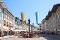 Belebte Fußgängerzone mit historischer Säule in der Mitte, umgeben von Bänken, Geschäften und bunten Fahnen an sonnigem Tag